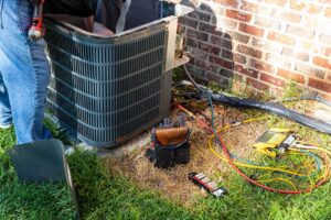Outdoor heat pump beside a home with a technician checking the indoor air handler and ductwork.