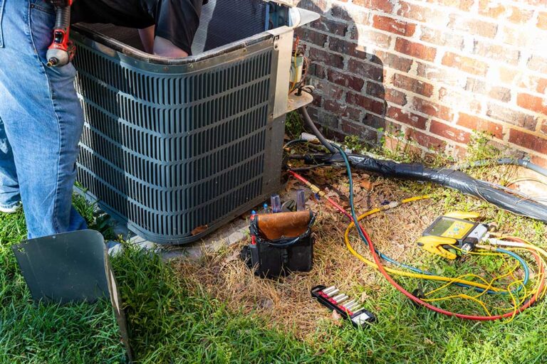 Outdoor heat pump beside a home with a technician checking the indoor air handler and ductwork.